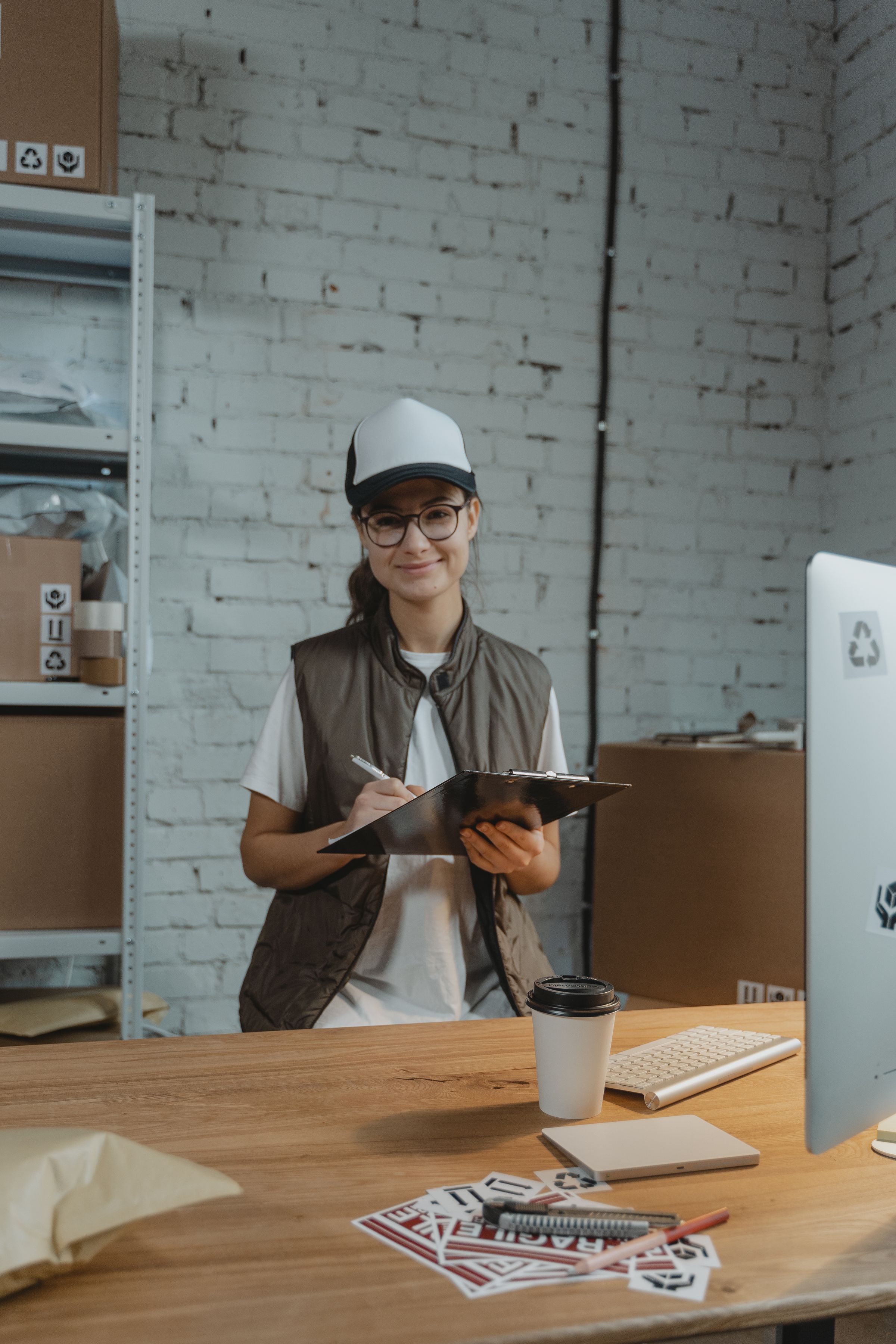 Smiling shipping-desk operator in a cap and glasses writing on a clipboard beside a monitor and shipping labels.